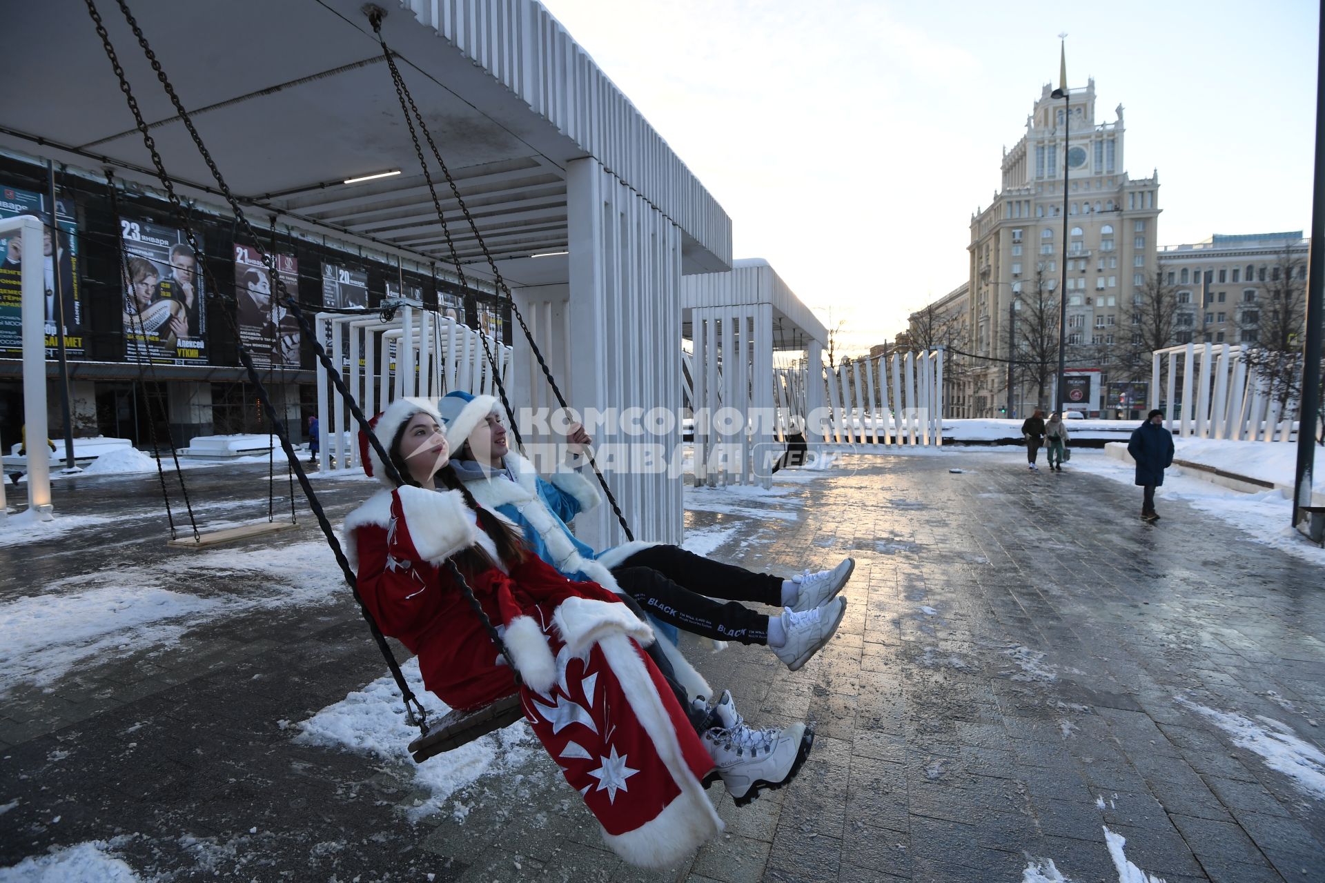 Москва.   Мужчина и женщина в костюмах деда Мороза и Снегурочки на качелях на Триумфальной площади.