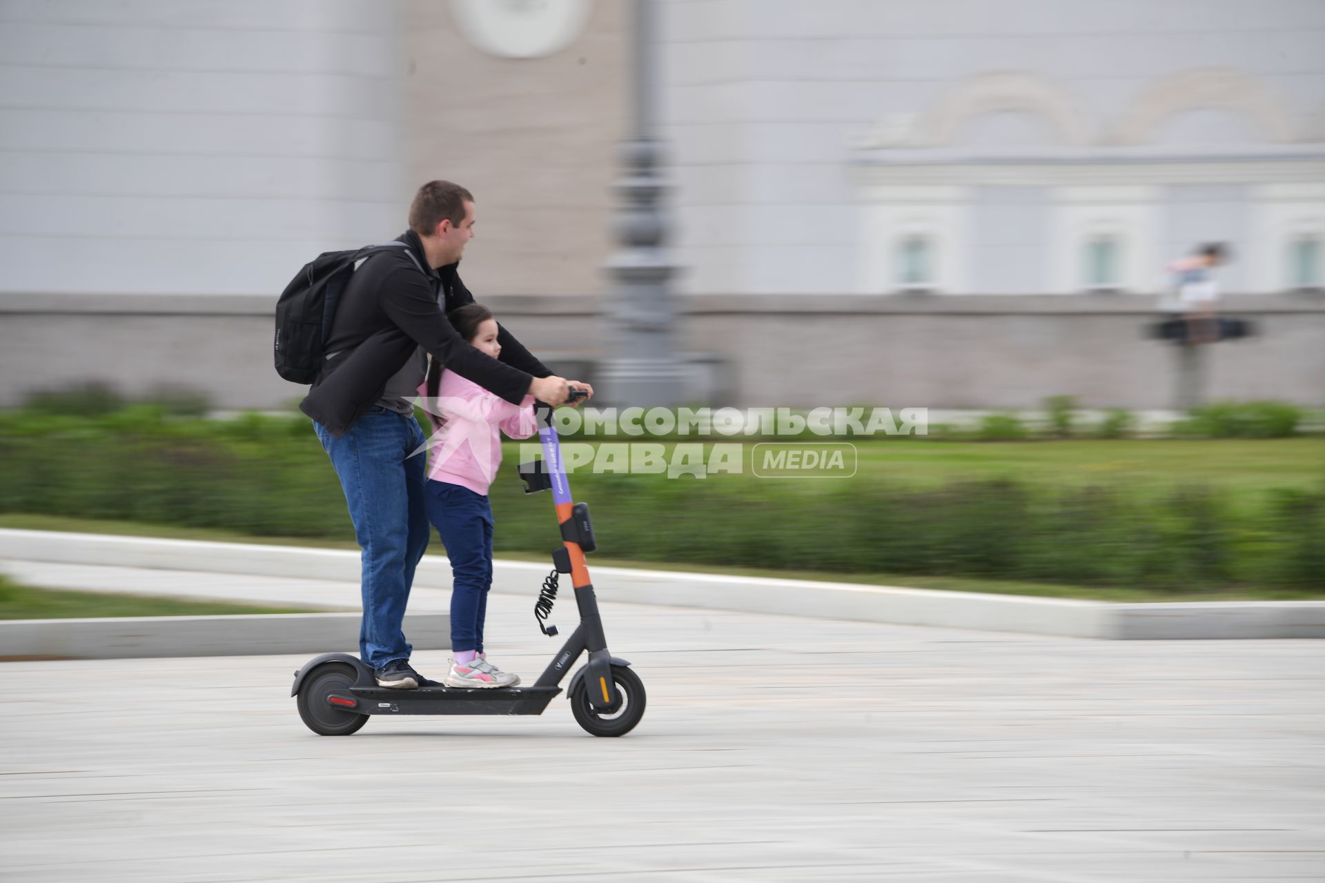 Москва.  Мужчина с девочкой едет на электросамокате  на ВДНХ.