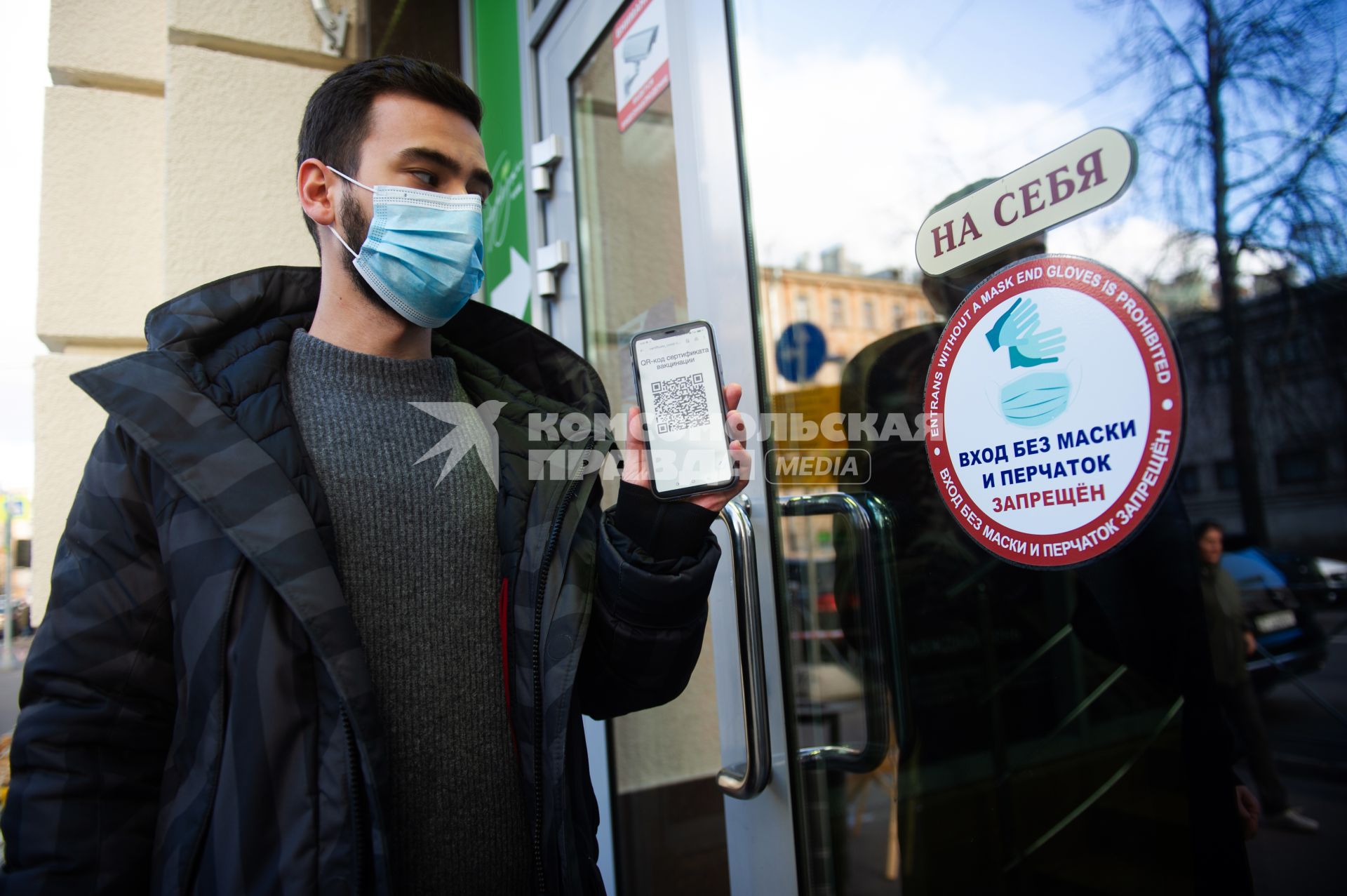 Санкт-Петербург. Юноша в маске и мобильным телефоном, на экране которого отображен QR-код, заходит в торговый центр. Наклейка на двери ТЦ `Вход без маски и перчаток запрещен`.