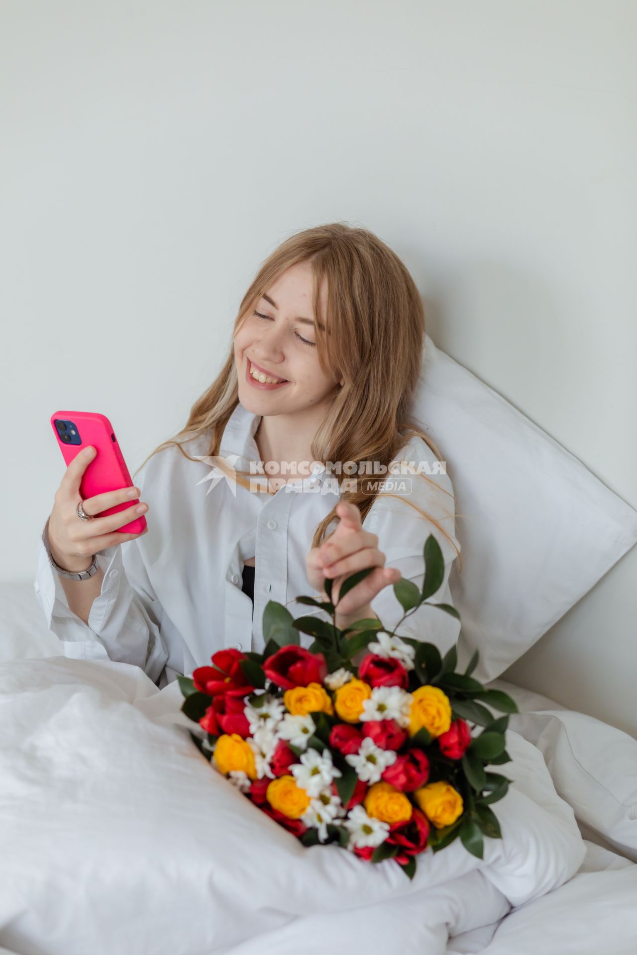 Красноярск. Девушка с мобильным телефоном и букетом цветов лежит в постели.