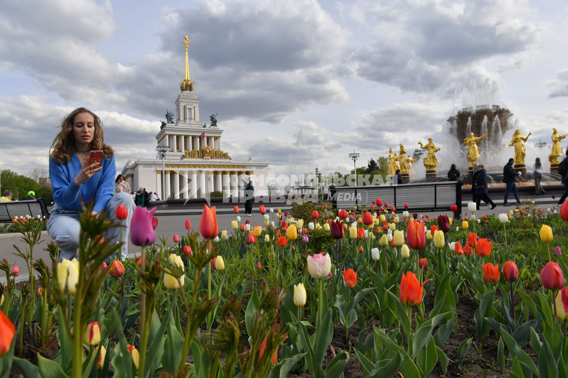 Москва. Девушка фотографирует на мобильный телефон тюльпаны рядом с павильоном #1 `Центральный` и фонтаном `Дружба народов` на ВДНХ.