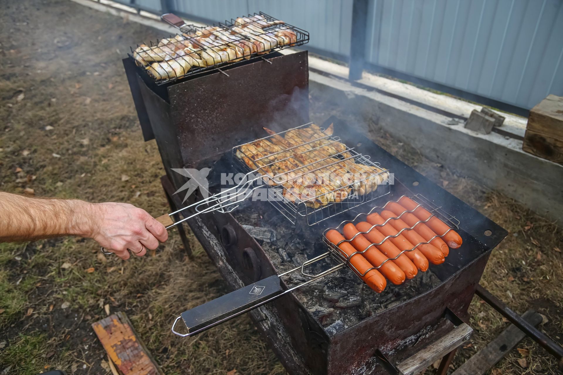 Красноярск. Приготовление куриных крылышек и сосисок на решетке на приусадебном участке.